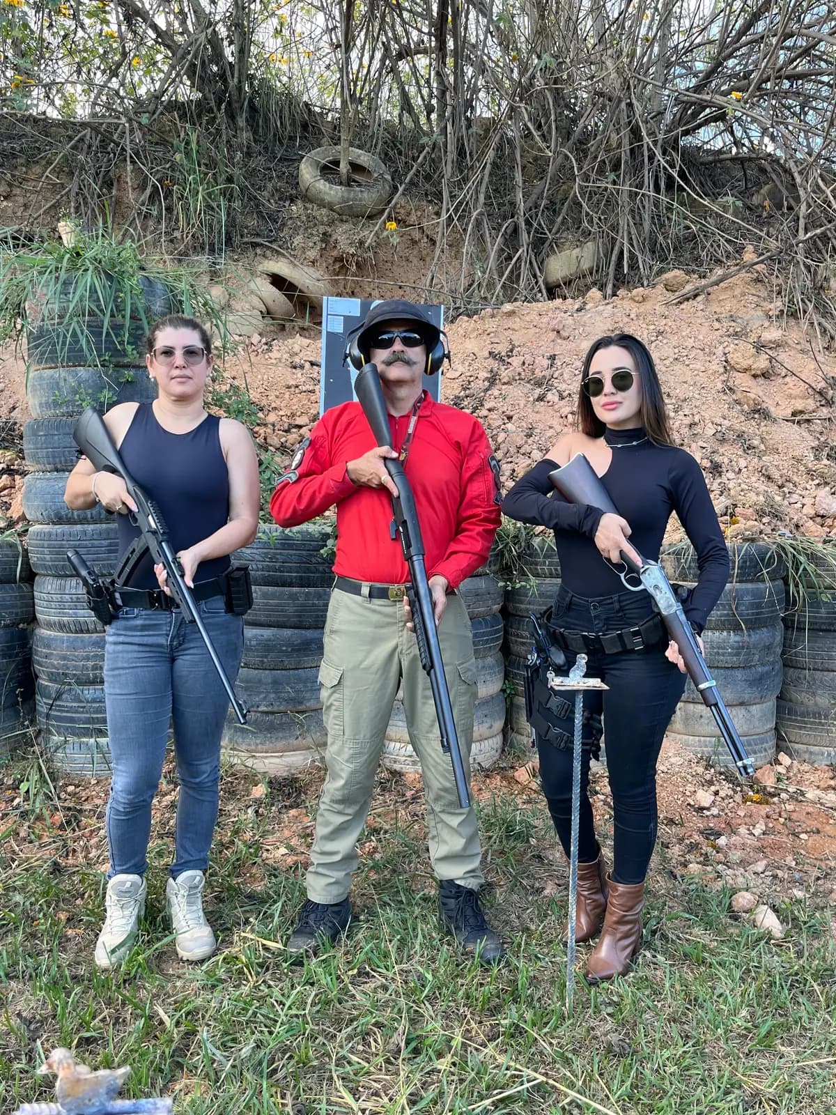 Três pessoas com armas longas em estande de tiro outdoor, com barreira de pneus ao fundo